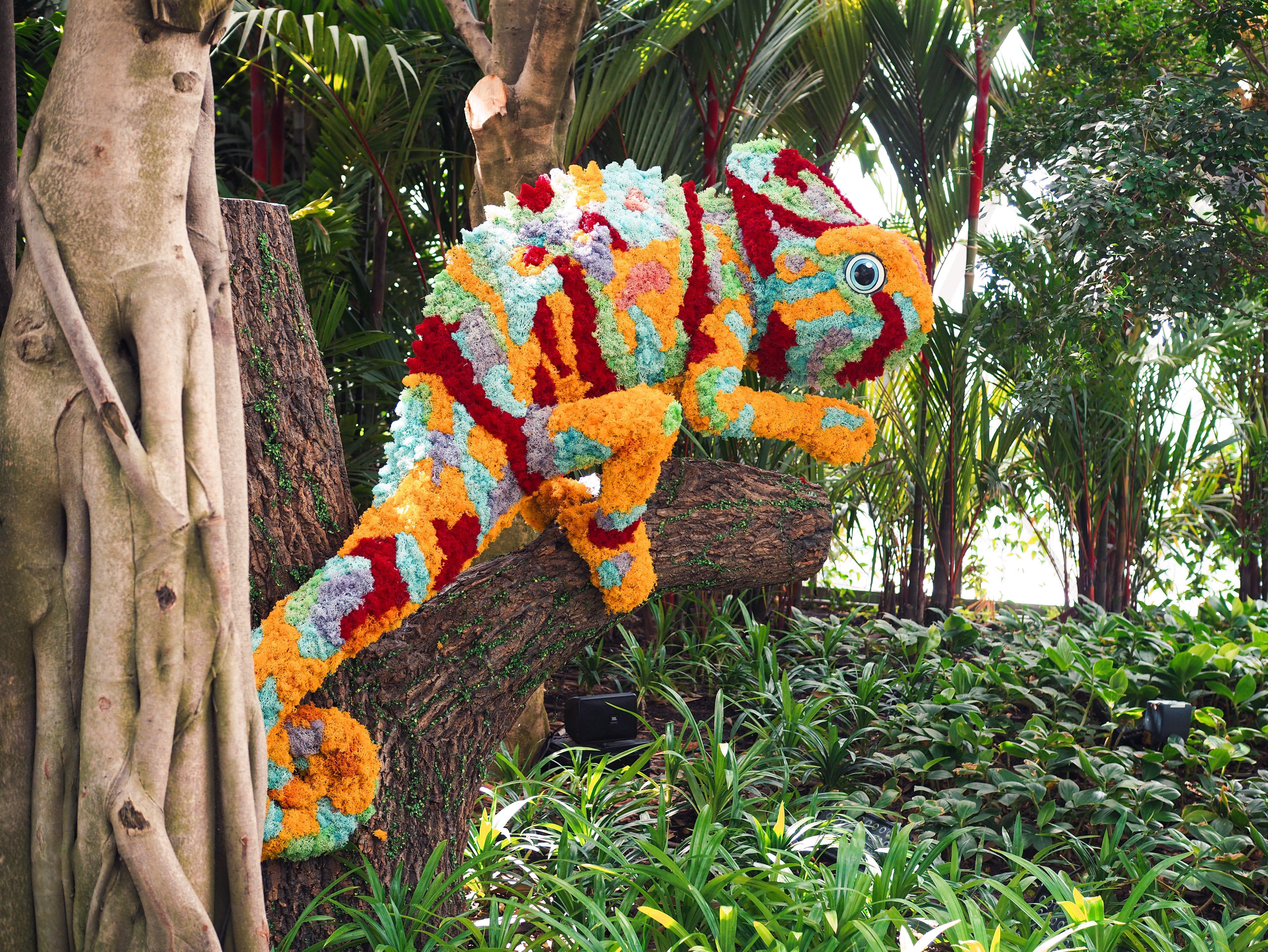 Crowne Plaza Changi Airport By Ihg Сингапур Экстерьер фото The photo shows a vibrant and colorful chameleon sculpture made from flowers and plant materials. The chameleon is positioned on a tree trunk, blending in with the lush greenery of its surroundings. The colors of the sculpture include shades of orang