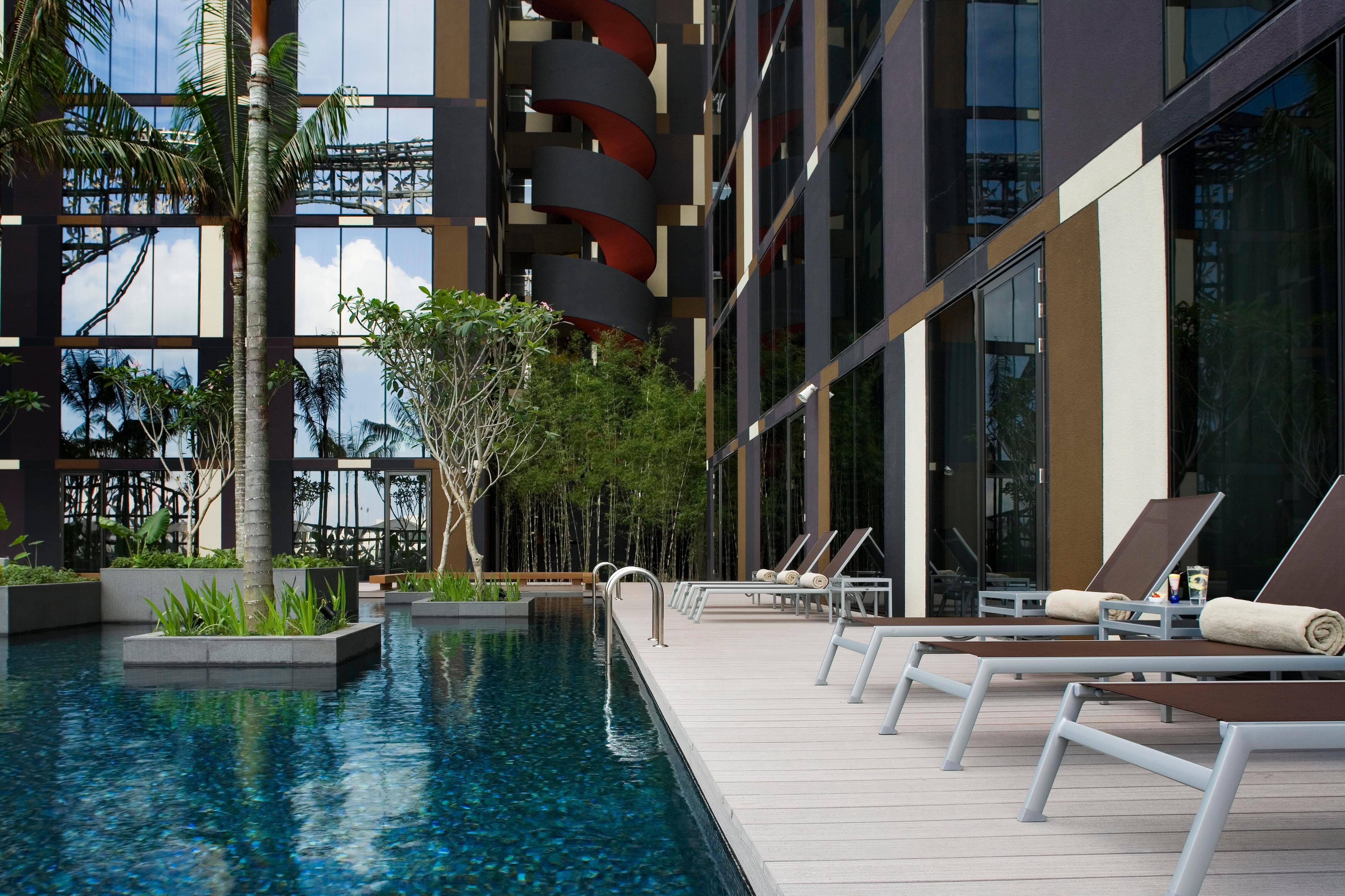 Crowne Plaza Changi Airport By Ihg Сингапур Удобства фото The photo shows a modern swimming pool area adjacent to a building. The pool is surrounded by sleek wooden decking and features a clear blue water surface. There are several lounge chairs positioned around the pool for relaxation. Lush greenery, incl