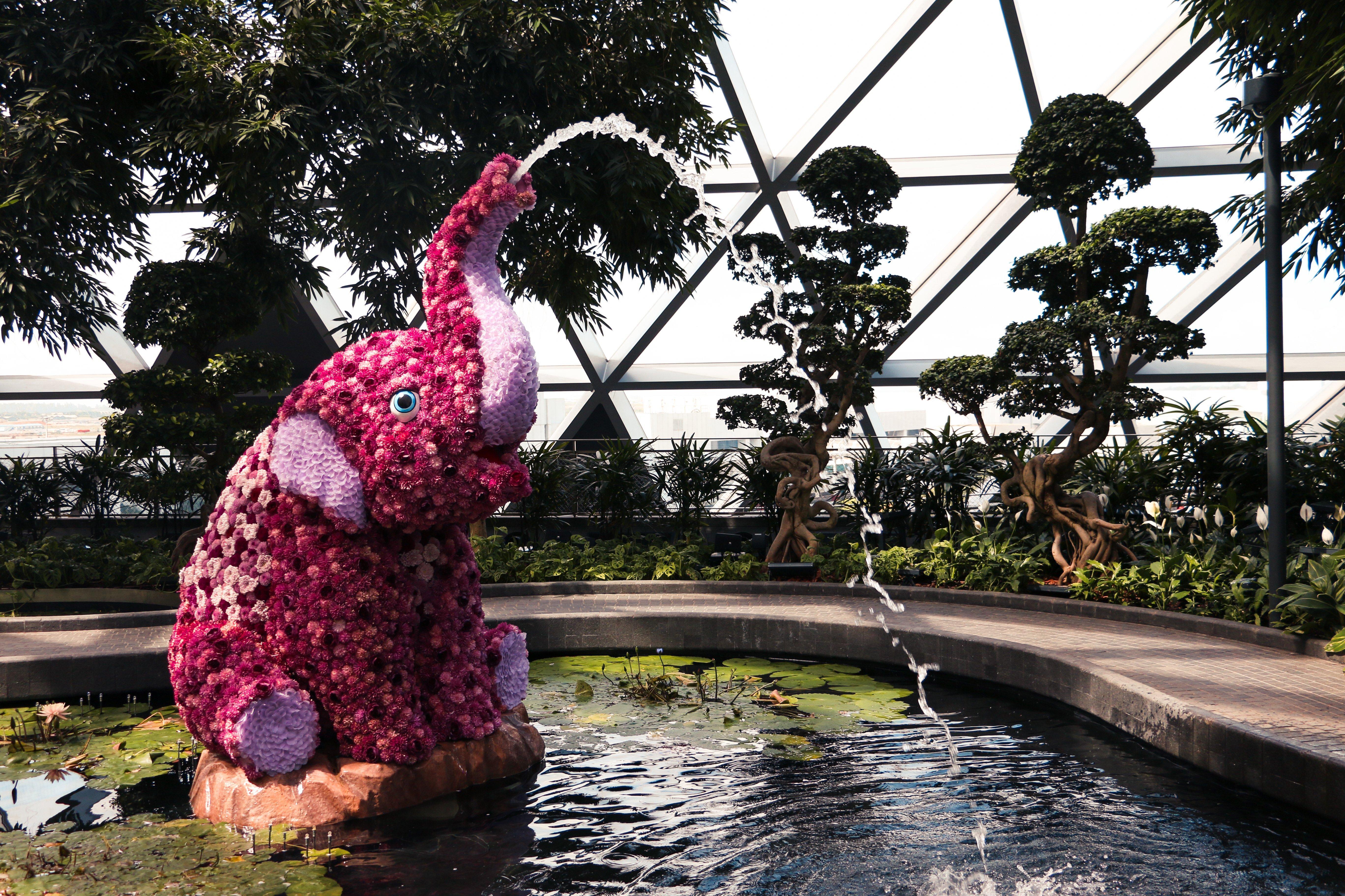 Crowne Plaza Changi Airport By Ihg Сингапур Экстерьер фото The photo depicts a decorative elephant sculpture made of flowers, positioned near a pond. The elephant is sitting on its hind legs, with its trunk raised high, creating a water fountain effect. Surrounding the elephant are lush green plants and tree