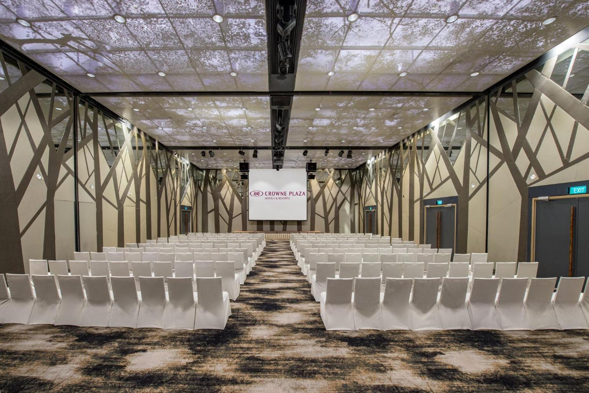 Crowne Plaza Changi Airport By Ihg Сингапур Экстерьер фото The photo shows a modern conference or event space. The room features rows of white chairs arranged in a theater-style layout, facing a large screen with a simple logo. The ceiling is designed to resemble a natural setting, with images that suggest t