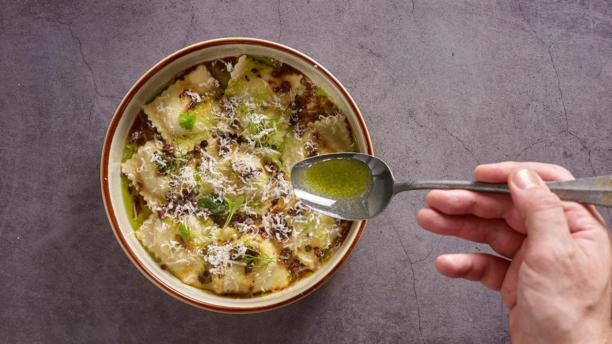 Crowne Plaza Changi Airport By Ihg Сингапур Экстерьер фото The photo shows a bowl of ravioli garnished with grated cheese and drizzled with olive oil. A hand is holding a spoon above the bowl, indicating the action of adding more oil or sauce. The dish appears delicious and is presented with an appealing aes