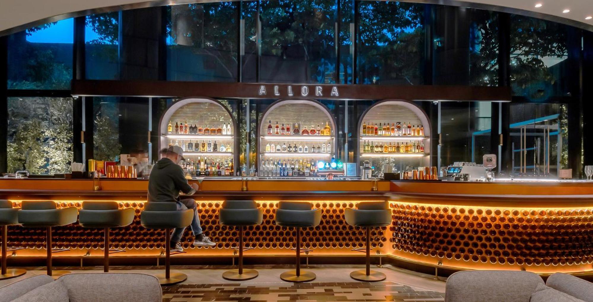 Crowne Plaza Changi Airport By Ihg Сингапур Экстерьер фото The photo depicts a modern bar interior with a sleek design. The bar is illuminated with warm lighting, highlighting a wide selection of alcohol displayed on shelves behind the counter. There are several high-backed stools at the bar, and a person is