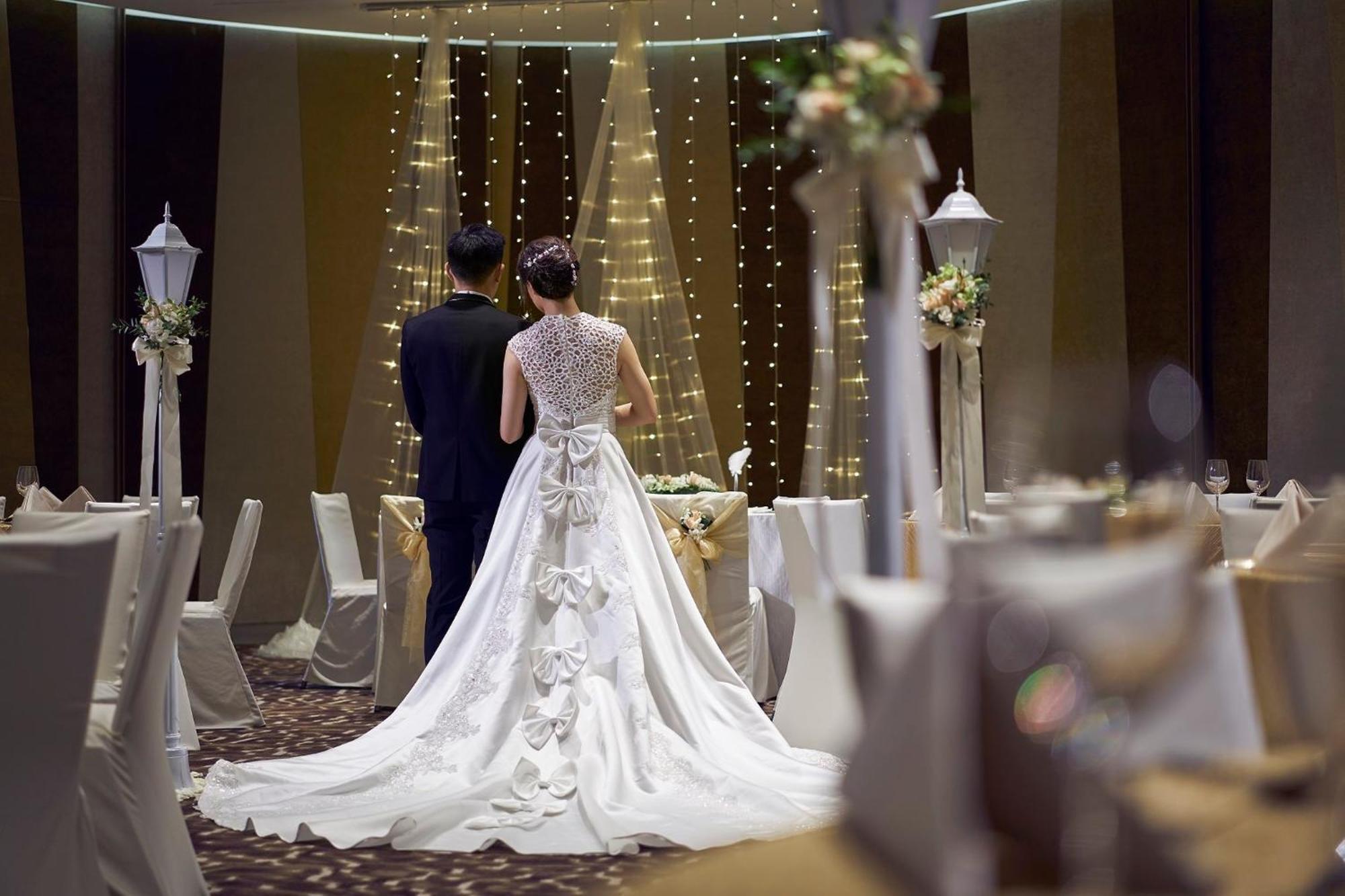 Crowne Plaza Changi Airport By Ihg Сингапур Экстерьер фото The photo features a couple at a wedding venue. The bride, wearing an elegant white gown with intricate details and a long train, is seen from behind. The groom stands beside her, dressed in a dark suit. The background showcases a beautifully decorat
