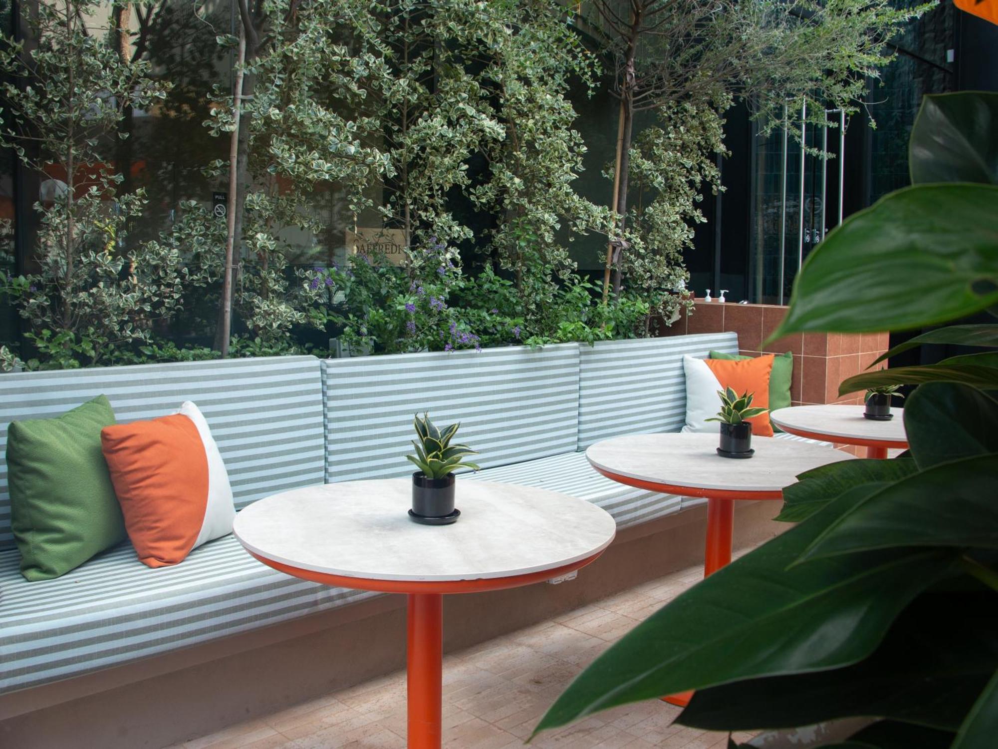 Crowne Plaza Changi Airport By Ihg Сингапур Экстерьер фото The photo shows a modern outdoor seating area. There is a long bench with a corrugated metal backrest, decorated with colorful cushions in green and orange. In front of the bench, there are circular tables with white marble tops and orange legs. Each