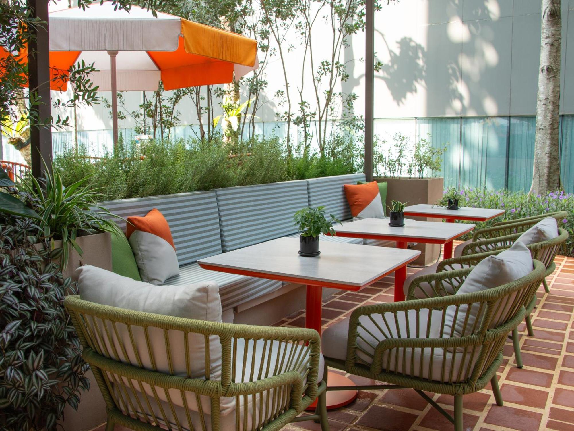 Crowne Plaza Changi Airport By Ihg Сингапур Экстерьер фото The photo shows a cozy outdoor seating area. There are several tables with red metal legs, each accompanied by comfortable chairs. The chairs have a woven design and are in shades of green. There are cushions in various colors on the chairs for added