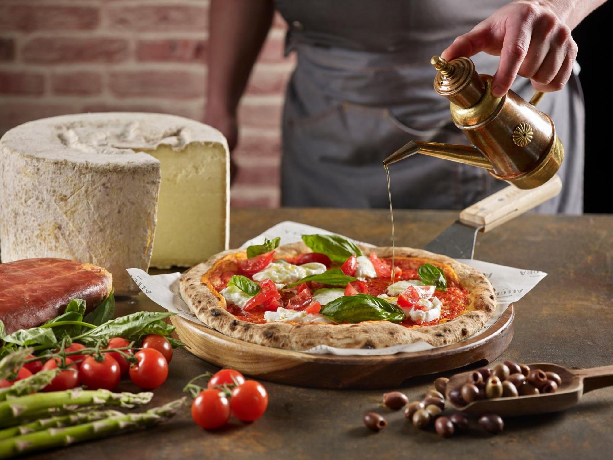Crowne Plaza Changi Airport By Ihg Сингапур Экстерьер фото The photo features a freshly made pizza being drizzled with oil. The pizza is topped with ingredients such as fresh basil and tomatoes, and has a golden, crispy crust. In the background, there is a block of cheese and a piece of ham. Surrounding the 