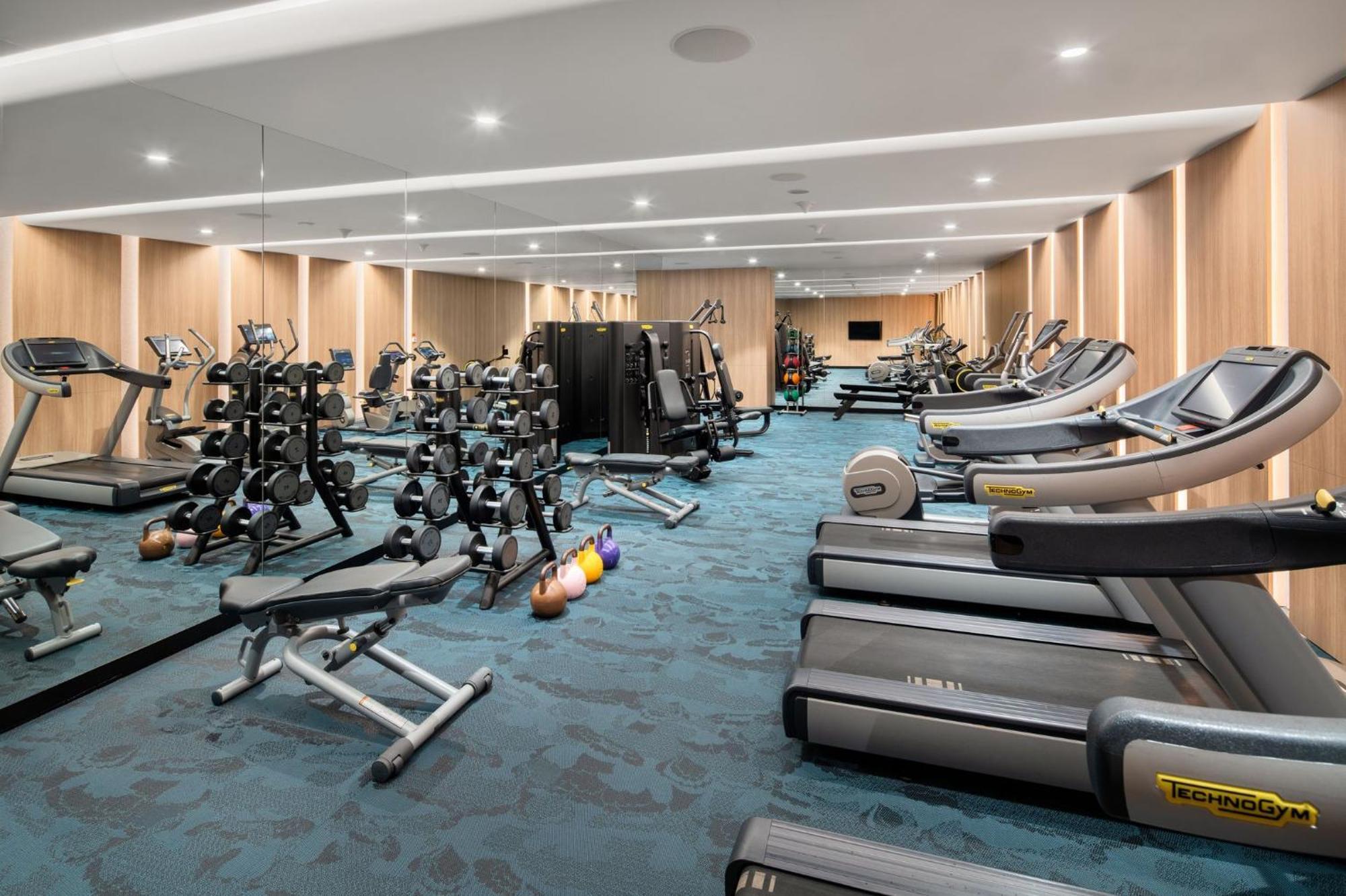 Crowne Plaza Changi Airport By Ihg Сингапур Экстерьер фото The photo shows a modern gym interior. It features various fitness equipment, including multiple treadmills, weightlifting machines, and a set of free weights on racks. The flooring has a blue carpet, and the walls are made of wood paneling, giving t