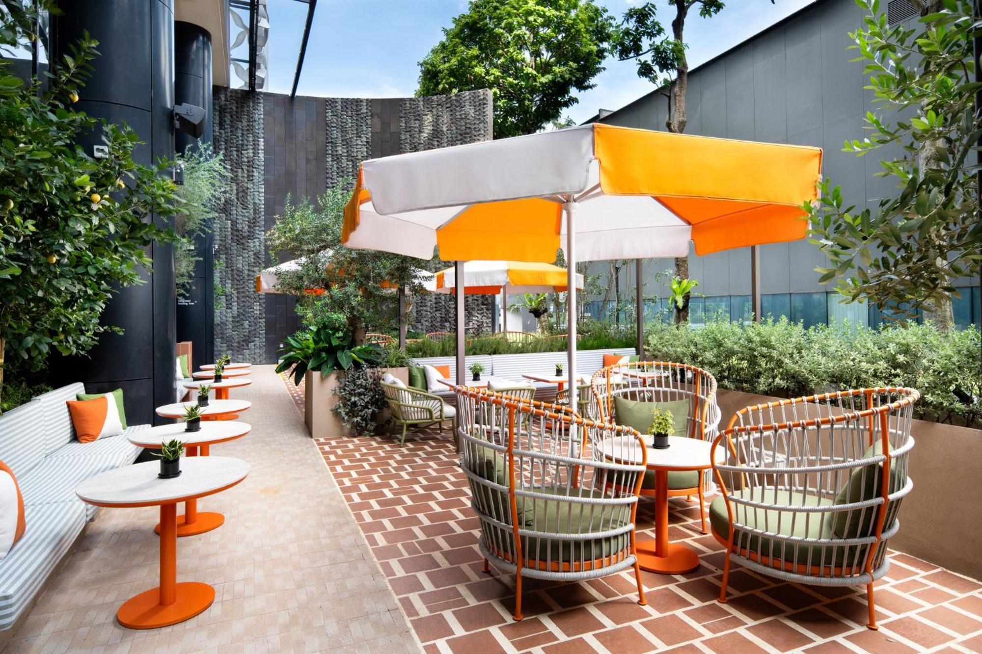 Crowne Plaza Changi Airport By Ihg Сингапур Экстерьер фото The photo depicts an outdoor seating area featuring several tables and chairs. The tables have orange bases and white tops, while the seating comprises rattan-style chairs with cushions. Each table is accompanied by a large umbrella with a white and 