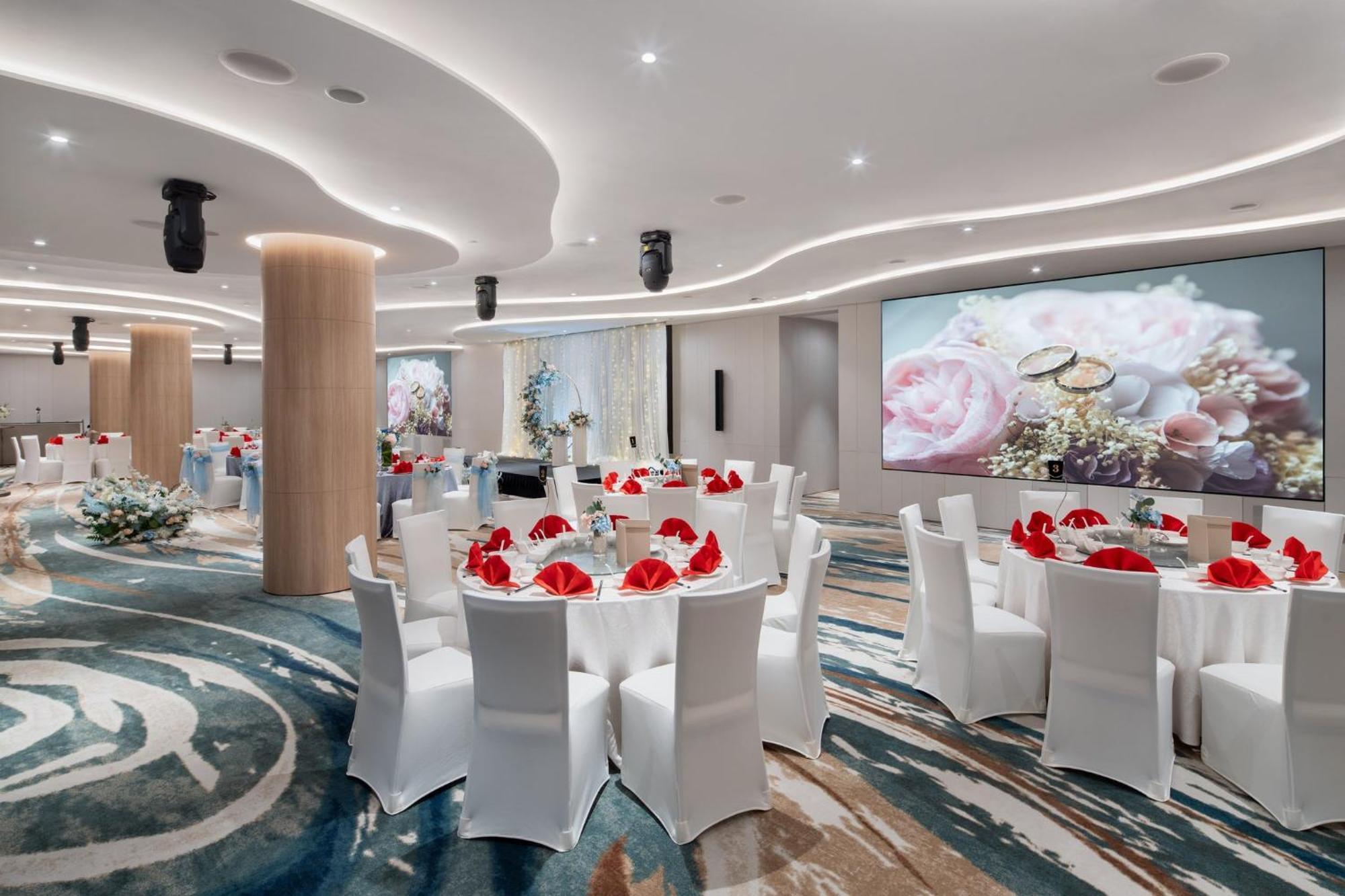 Crowne Plaza Changi Airport By Ihg Сингапур Экстерьер фото The photo depicts an elegantly designed banquet or event hall. It features round dining tables covered with white tablecloths, each adorned with red napkins. The chairs are also white, creating a cohesive color scheme. The room has modern lighting an