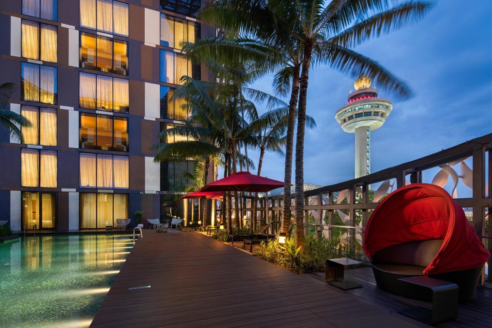 Crowne Plaza Changi Airport By Ihg Сингапур Экстерьер фото The image shows a modern hotel property situated near a swimming pool. The hotel features large windows with warm lighting, and it is surrounded by palm trees. There are red umbrellas providing shade, and a unique red seating area is visible near the