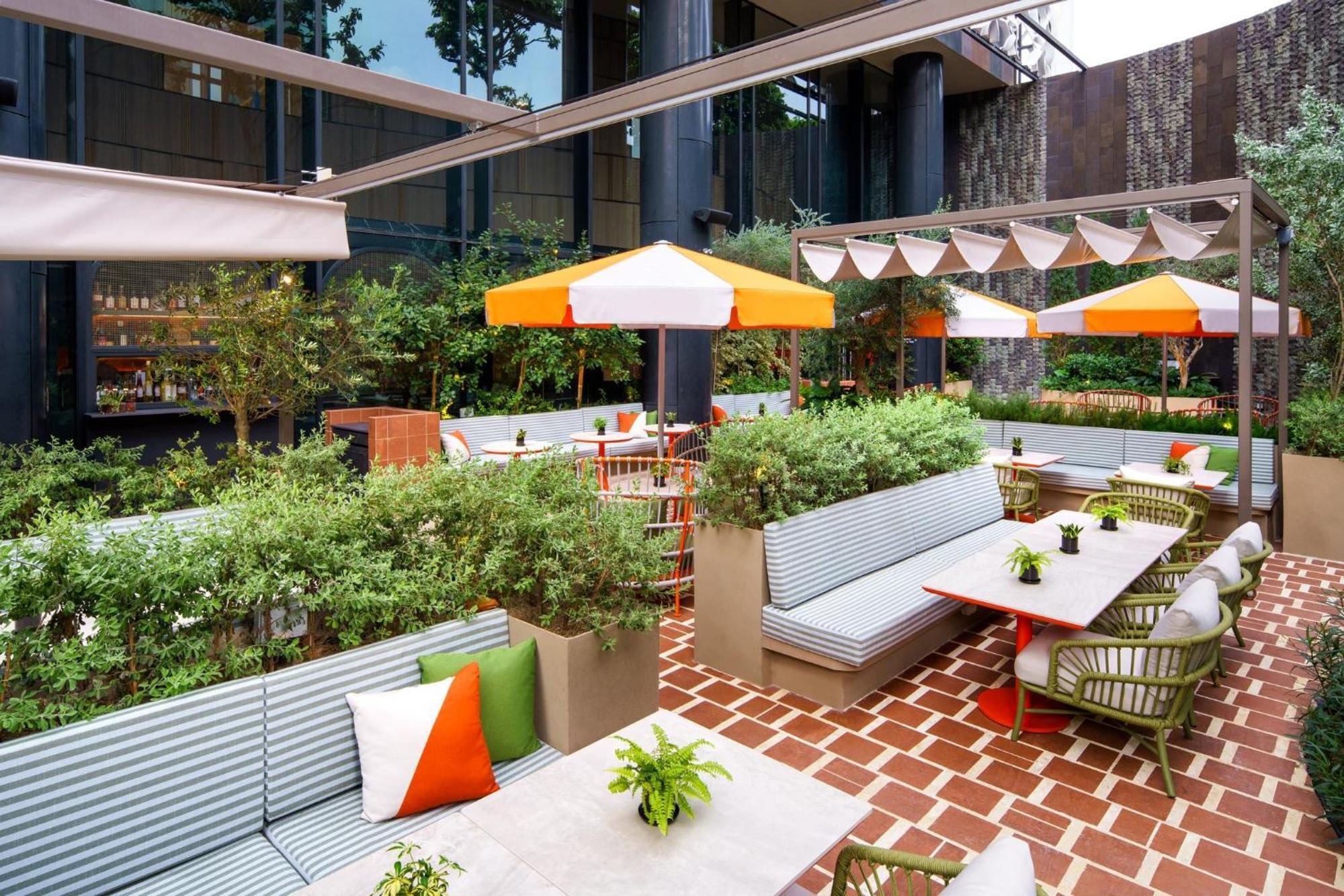 Crowne Plaza Changi Airport By Ihg Сингапур Экстерьер фото The photo depicts a stylish outdoor seating area in a modern setting. It features several light gray benches with colorful cushions, arranged around tables. The space is adorned with green plants and small potted flowers, adding a touch of nature. Th